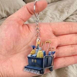 May include: A blue cleaning cart keychain with various cleaning supplies. The cart has a brush, spray bottles, and a bottle of cleaner. The keychain is attached to a silver chain and ring.