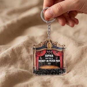 May include: A clear acrylic keychain featuring a theater stage design. The stage has red curtains, a wooden floor, and black seating. Text reads "SOPHIA starring as WENDY IN PETER PAN 2026". The keychain is attached to a silver keyring.