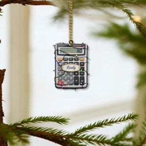 May include: A calculator Christmas ornament with the name "Emily" on a gold label. The calculator is wrapped in a string of colorful lights and hangs from a gold cord. The display shows "180".