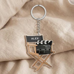 May include: A clear acrylic keychain featuring a director's chair and film clapperboard design. The chair has a black back with the name "ALEX" printed on it. The clapperboard has text that reads "PRODUCTION, DIRECTOR, CAMERA, DATE, SCENE, TAKE".