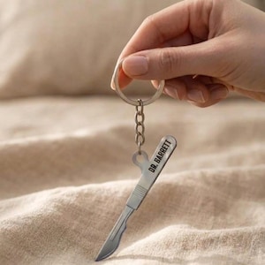 May include: A silver-colored scalpel keychain with the text "DR. BARRETT" engraved on the handle. The keychain is attached to a silver key ring and chain. The image is set against a neutral background.