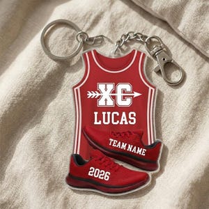 May include: A red acrylic keychain featuring a cross country jersey and running shoes. The jersey has "XC" and "LUCAS" in white. The shoes have "TEAM NAME" and "2026" in white. The keychain has a silver keyring and clasp.