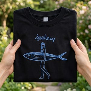 May include: Black t-shirt featuring a blue illustration of a woman carrying a fish, with the word "Sardiney" above. The design is a humorous take on a surfer girl. The shirt is folded, displaying the design.