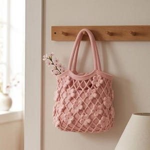 May include: A light pink, crocheted tote bag with a floral design hangs on a wooden peg rack. The bag has two handles and is filled with a sprig of white flowers. The bag is hanging on a light beige wall.