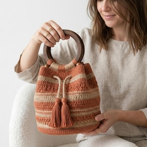 May include: A handcrafted crochet bucket bag with a wooden handle. The bag has horizontal stripes in orange and beige, with matching tassels. The bag is held by a person.