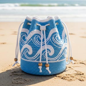 May include: A blue and white crochet bucket bag with a wave pattern. The bag has a drawstring closure with white cords and wooden beads. The bag is sitting on a sandy beach with the ocean in the background.