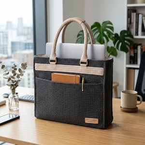 May include: A dark gray tote bag with a beige stripe and handles. The bag has a front pocket with a notebook and pen. A laptop is inside the bag. The handles are beige leather. The bag is on a wooden desk.