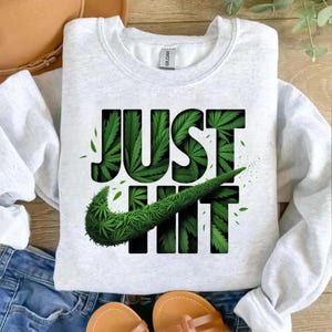 May include: A pale grey sweatshirt featuring the text "JUST HIT" in black, with a Nike swoosh made of green cannabis leaves. The sweatshirt is displayed on a wooden surface with blue denim jeans and sandals.