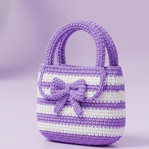 May include: A handmade crochet handbag in shades of purple and white. The bag features a striped pattern and a decorative bow on the front. It has a rounded handle and a flap closure. The bag is designed with a soft, textured appearance.