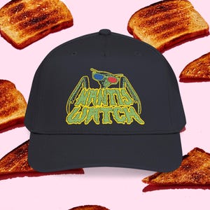 May include: A navy blue baseball cap with the text "Mantis Watch" in yellow and green. The graphic features a green insect wearing 3D glasses. The background is a light pink with slices of toasted bread.
