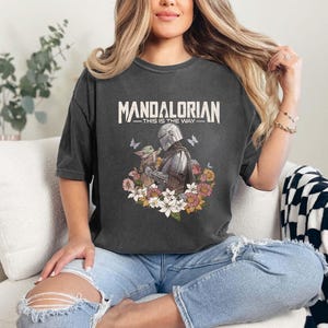 May include: Dark gray t-shirt featuring the text "MANDALORIAN" above an illustration of the Mandalorian and Grogu surrounded by flowers and butterflies. The phrase "THIS IS THE WAY" is below the title.