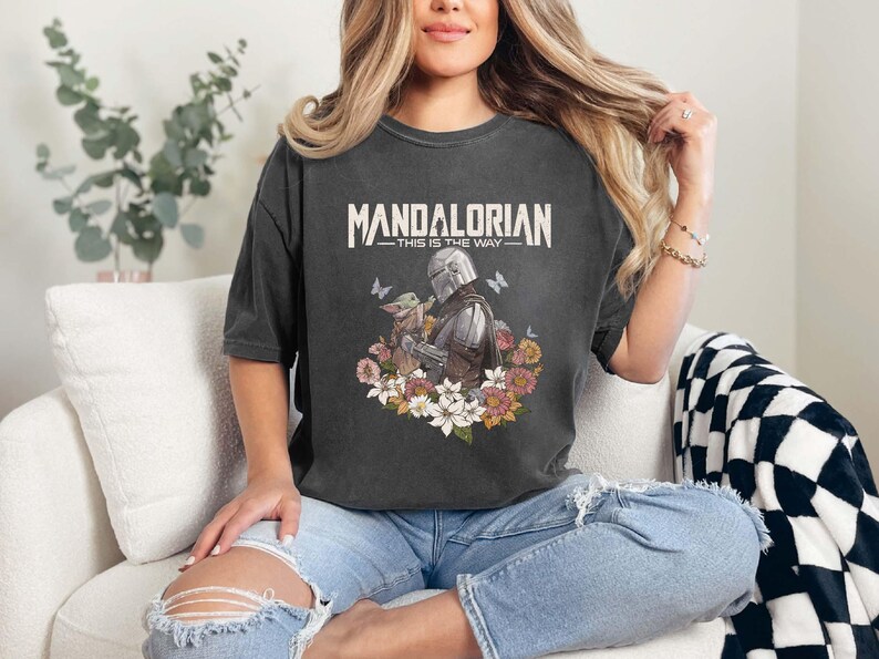May include: Dark gray t-shirt featuring the text "MANDALORIAN" above an illustration of the Mandalorian and Grogu surrounded by flowers and butterflies. The phrase "THIS IS THE WAY" is below the title.