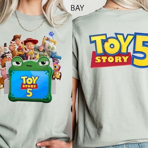 May include: A light sage green t-shirt featuring a graphic of Toy Story characters surrounding a green frog-shaped frame with the text Toy Story 5 on the front, and the text Toy Story 5 in large yellow and blue letters on the back.
