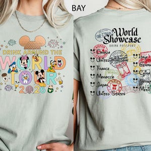 May include: A pale green t-shirt with a vibrant graphic of Disney characters and the text "DRINK AROUND THE WORLD TOUR 2026" on the front. The back features passport stamps with the words "World Showcase Drink Passport".