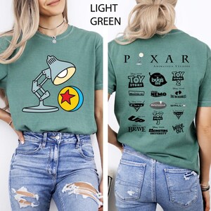 May include: Light green t-shirt featuring a cartoon image of a desk lamp and a yellow ball with a red star. The back of the shirt displays the Pixar Animation Studios logo and various movie titles.