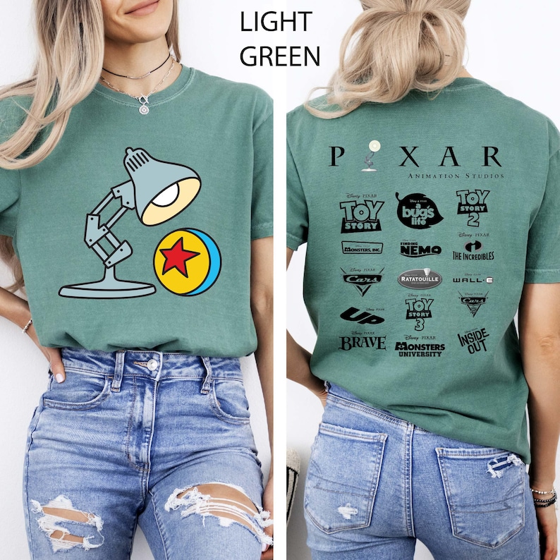 May include: Light green t-shirt featuring a cartoon image of a desk lamp and a yellow ball with a red star. The back of the shirt displays the Pixar Animation Studios logo and various movie titles.