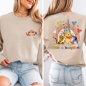 Op de afbeelding: Beige sweatshirt met een kleine afbeelding van Winnie de Poeh, Teigetje en Knorretje op de voorkant. De achterkant heeft een groter ontwerp met de personages, een kasteel, een regenboog en de tekst "Walt Disney World".