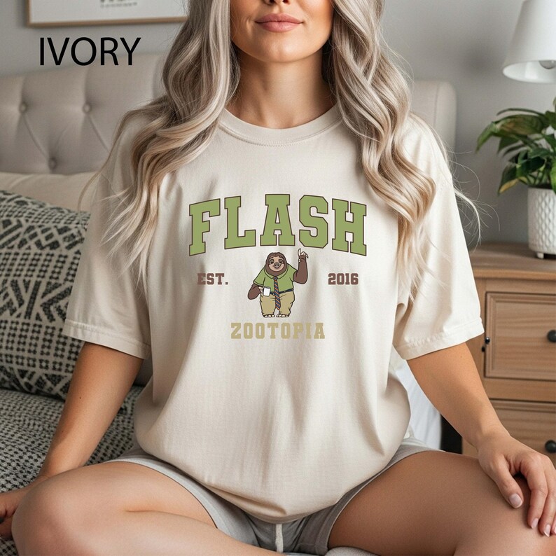 May include: Ivory-colored t-shirt with the word "FLASH" in green block letters. Below the word "FLASH" is a cartoon sloth character with the text "EST. 2016 ZOOTOPIA". The shirt is a casual style.