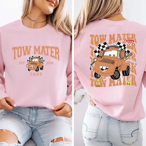 Peut inclure: Sweat-shirt rose clair avec une image de dessin anim&eacute; de Martin de Cars. Le devant affiche "Tow Mater" au-dessus de l'image, avec "Est. 2006" et "Cars" en dessous. Le dos pr&eacute;sente une image plus grande de Martin.