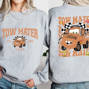 Peut inclure: Un sweat-shirt gris clair avec une image de dessin anim&eacute; de Martin la d&eacute;panneuse du film Cars. Le devant affiche "TOW MATER" au-dessus de l'image, avec "EST. 2006" et "CARS" en dessous. Le dos pr&eacute;sente Martin avec des drapeaux &agrave; damier.