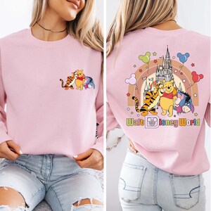 Op de afbeelding: Een lichtroze sweatshirt met een cartoonafbeelding van Winnie de Poeh, Teigetje en Iejoor. De voorkant heeft de karakters samen, en de achterkant heeft de karakters met een regenboog, kasteel en de tekst "Walt Disney World".