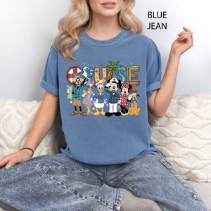 May include: Blue t-shirt with the word "CRUISE" in colorful letters and a graphic of Disney characters. The characters include Mickey Mouse, Minnie Mouse, Donald Duck, and Goofy. The shirt is a light blue color.