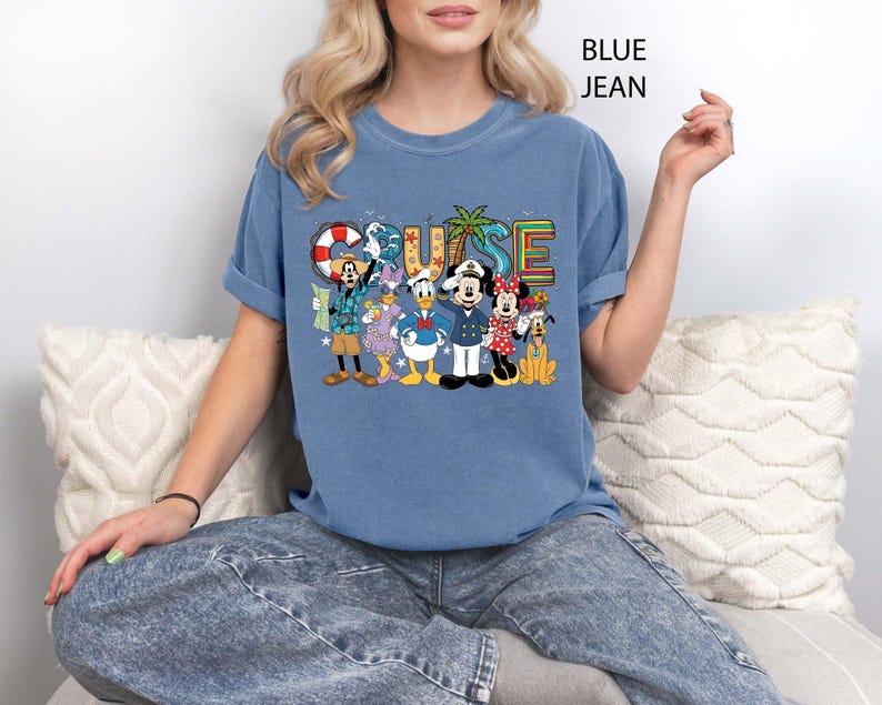 May include: Blue t-shirt with the word "CRUISE" in colorful letters and a graphic of Disney characters. The characters include Mickey Mouse, Minnie Mouse, Donald Duck, and Goofy. The shirt is a light blue color.