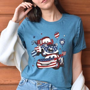 May include: A heather blue t-shirt featuring a graphic print of two cartoon cars decorated with red, white, and blue American flag patterns. The design includes balloons, one marked with the number 95, and small stars scattered around the vehicles.