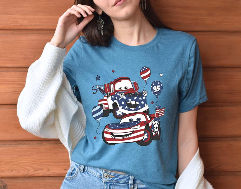May include: A heather blue t-shirt featuring a graphic print of two cartoon cars decorated with red, white, and blue American flag patterns. The design includes balloons, one marked with the number 95, and small stars scattered around the vehicles.