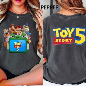 May include: A charcoal grey t-shirt featuring a front graphic of Toy Story characters surrounding a green frog-shaped frame with the text Toy Story 5. The back of the shirt displays the large yellow and blue text Toy Story 5.
