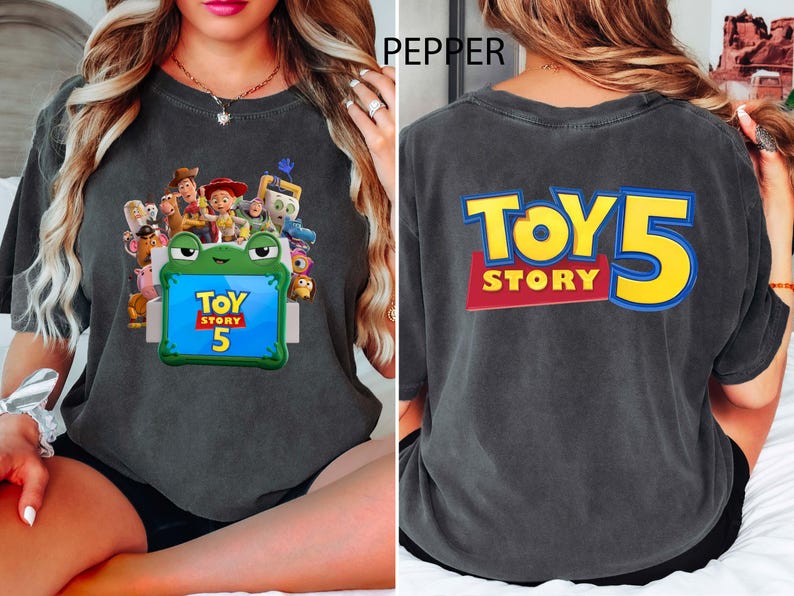 May include: A charcoal grey t-shirt featuring a front graphic of Toy Story characters surrounding a green frog-shaped frame with the text Toy Story 5. The back of the shirt displays the large yellow and blue text Toy Story 5.