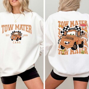 Peut inclure: Sweat-shirt blanc avec une image de dessin anim&eacute; de Martin la d&eacute;panneuse du film Cars. Le design avant comprend le texte "TOW MATER EST. 2006 CARS". Le design arri&egrave;re montre Martin avec des drapeaux &agrave; damiers.
