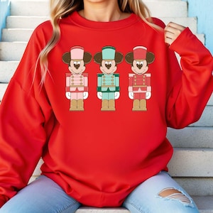 May include: A red sweatshirt featuring a graphic of three toy soldier-style Mickey Mouse characters. Each character is dressed in a different color uniform: pink, teal, and red. The design has a cartoonish style, with details like buttons and hats.