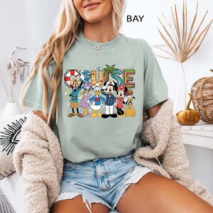 May include: A light green t-shirt with the word "CRUISE" in colorful letters and a graphic of Disney characters. The characters include Mickey Mouse, Minnie Mouse, Donald Duck, and Goofy. The shirt is a comfortable, casual style.