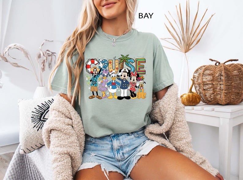 May include: A light green t-shirt with the word "CRUISE" in colorful letters and a graphic of Disney characters. The characters include Mickey Mouse, Minnie Mouse, Donald Duck, and Goofy. The shirt is a comfortable, casual style.