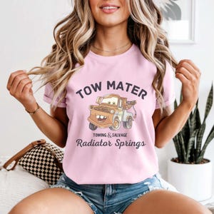 Peut inclure: T-shirt rose clair avec une image de dessin anim&eacute; de Tow Mater du film Cars. Le texte "TOW MATER" est au-dessus de l'image, et "TOWING & SALVAGE" et "Radiator Springs" sont en dessous.