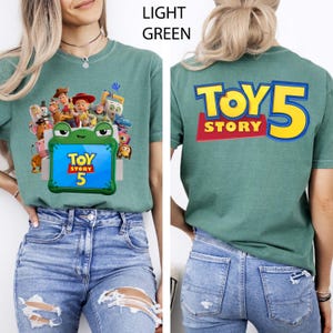 May include: A light green t-shirt featuring a front graphic of Toy Story characters surrounding a green frog-shaped frame with the text Toy Story 5 inside. The back of the shirt displays the large yellow and blue text Toy Story 5.