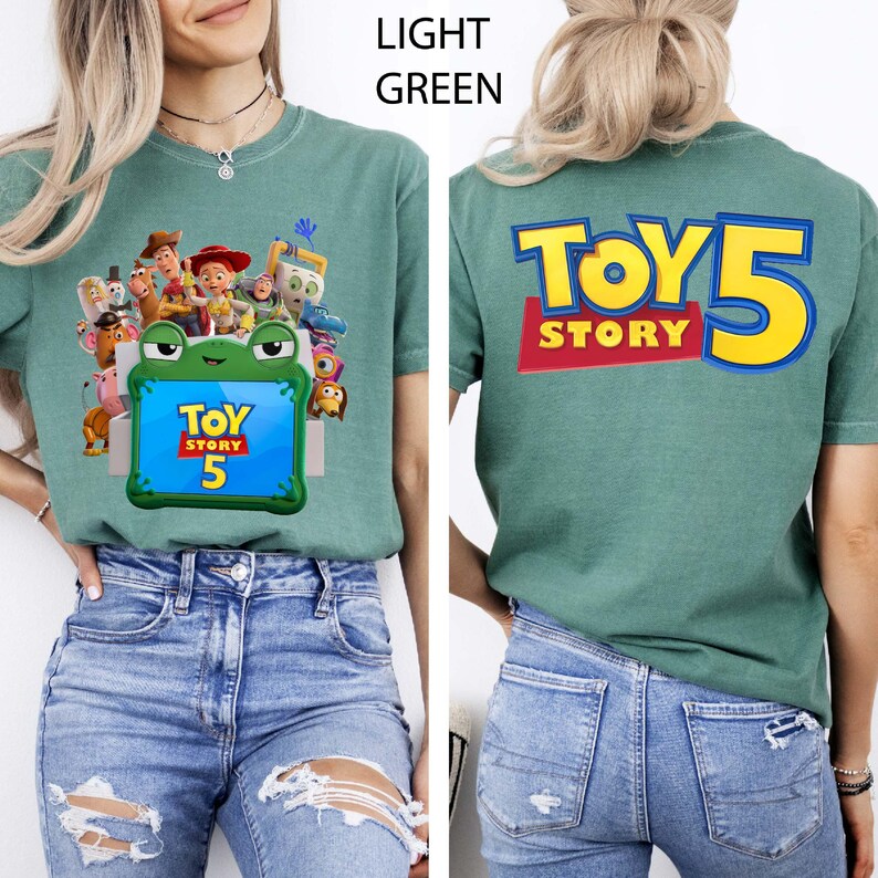 May include: A light green t-shirt featuring a front graphic of Toy Story characters surrounding a green frog-shaped frame with the text Toy Story 5 inside. The back of the shirt displays the large yellow and blue text Toy Story 5.