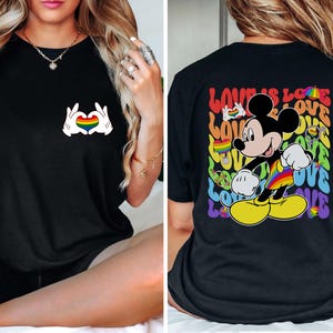 May include: Black t-shirt featuring a Mickey Mouse design with a rainbow-colored outfit and the words "LOVE IS LOVE". The front of the shirt has a heart made of hands with a rainbow heart in the middle.