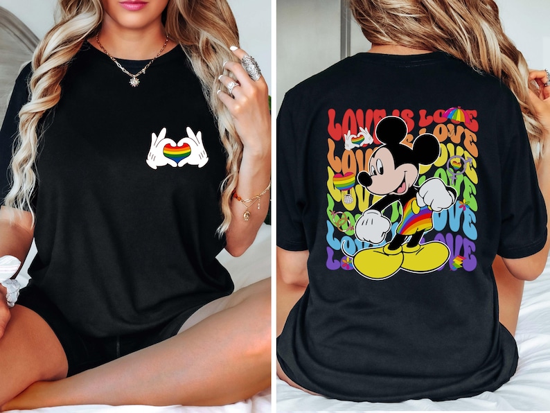 May include: Black t-shirt featuring a Mickey Mouse design with a rainbow-colored outfit and the words "LOVE IS LOVE". The front of the shirt has a heart made of hands with a rainbow heart in the middle.