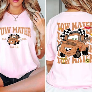 Puede incluir: Camiseta rosa claro con una imagen de dibujos animados de Mate de la pel&iacute;cula Cars. La parte delantera muestra "Tow Mater Cars Est. 2006". La parte trasera muestra una imagen m&aacute;s grande de Mate con banderas a cuadros y el texto "Tow Mater".
