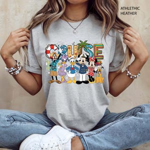 May include: Athletic heather gray t-shirt featuring the word "CRUISE" in colorful letters with cartoon characters. The characters include Mickey Mouse, Minnie Mouse, Donald Duck, Goofy, and others, all dressed in cruise attire.