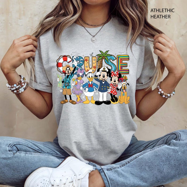 May include: Athletic heather gray t-shirt featuring the word "CRUISE" in colorful letters with cartoon characters. The characters include Mickey Mouse, Minnie Mouse, Donald Duck, Goofy, and others, all dressed in cruise attire.