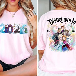 K&ouml;nnte beinhalten: Hellrosa T-Shirt mit der Aufschrift "2026" und Frozen-Figuren. Auf der R&uuml;ckseite steht "Disneyworld Est 2013" mit einer farbenfrohen Grafik der Charaktere. Ein lustiges Design f&uuml;r Disney-Fans.