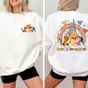 Op de afbeelding: Witte sweatshirt met een kleurrijke afbeelding van Winnie de Poeh, Teigetje, Iejoor en een kasteel. De voorkant heeft de karakters en de achterkant heeft de karakters, een regenboog en de tekst "Walt Disney World".