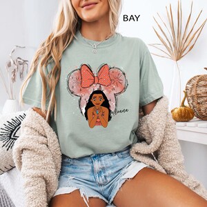 Pu&ograve; includere: T-shirt verde chiaro con un'illustrazione a cartoni animati di Moana all'interno di una silhouette di Topolino con un fiocco rosa. La parola "Moana" &egrave; scritta in corsivo sotto il personaggio. La parola "BAY" &egrave; stampata in alto.
