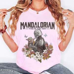 May include: Pink t-shirt featuring a graphic of The Mandalorian and Grogu surrounded by flowers and butterflies. The text "MANDALORIAN - THIS IS THE WAY" is printed across the top.