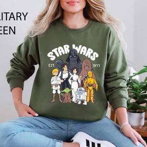 Puede incluir: Sudadera verde militar con un diseño de Star Wars, con el texto "STAR WARS" y "EST. 1977". El diseño presenta ilustraciones de dibujos animados de personajes como Darth Vader, Luke Skywalker y C-3PO. La sudadera es de cuello redondo.