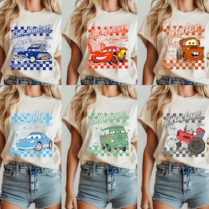 May include: A collection of six beige t-shirts featuring cartoon car designs. Each shirt displays a different character: Doc Hudson, McQueen, Mater, Sally, Fillmore, and Tractors. The designs include text and checkered patterns.