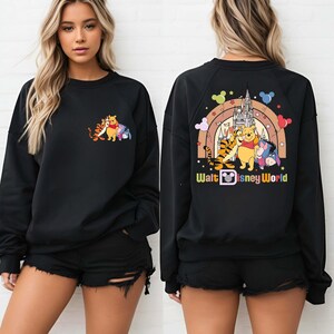 Op de afbeelding: Zwart sweatshirt met een kleurrijke afbeelding van Winnie de Poeh, Teigetje, Iejoor en een kasteel met een regenboog. De tekst "Walt Disney World" staat eronder. Het sweatshirt wordt door een persoon gedragen.